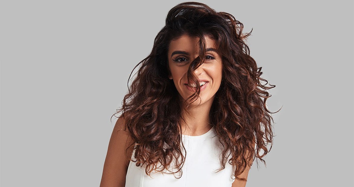 Morena iluminada com cachos médios e um sorriso natural, com o cabelo caindo no rosto